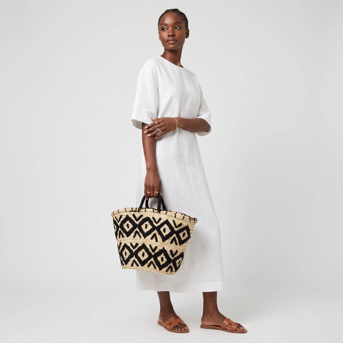 A woman modeling a white midi dress and brown leather sandals, holding a woven straw tote bag with a black geometric pattern against a light gray studio background.