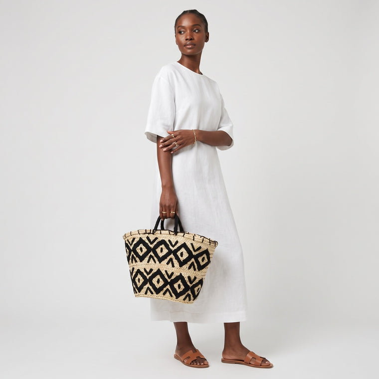 A woman modeling a white midi dress and brown leather sandals, holding a woven straw tote bag with a black geometric pattern against a light gray studio background.