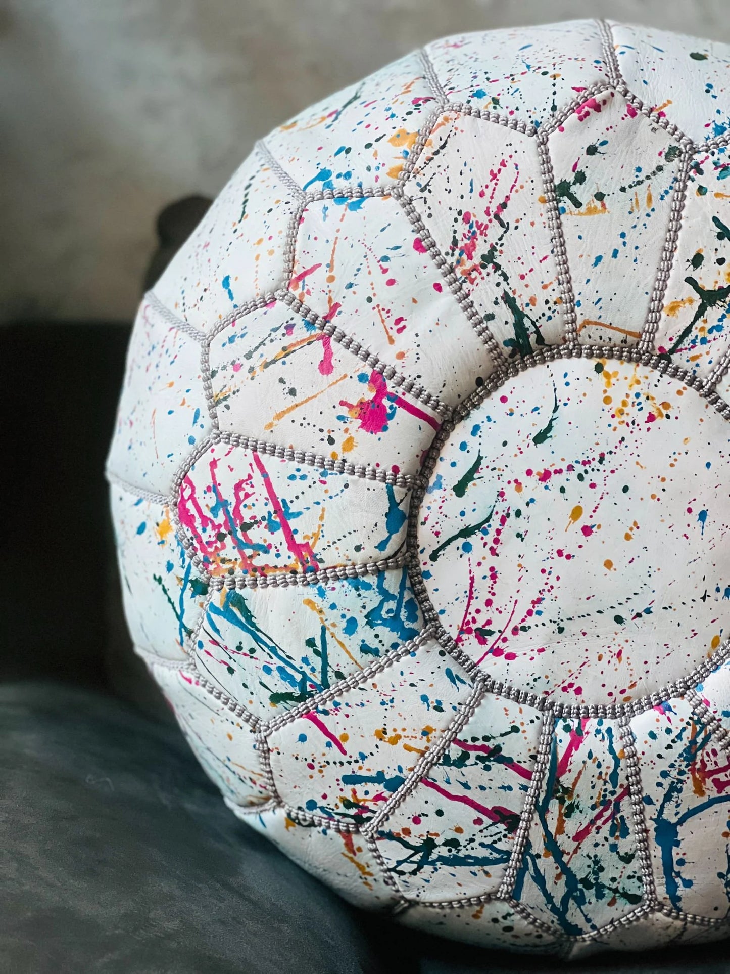 A close-up of a round pouf or cushion with a white base covered in colorful paint splatters and stitched geometric panels.