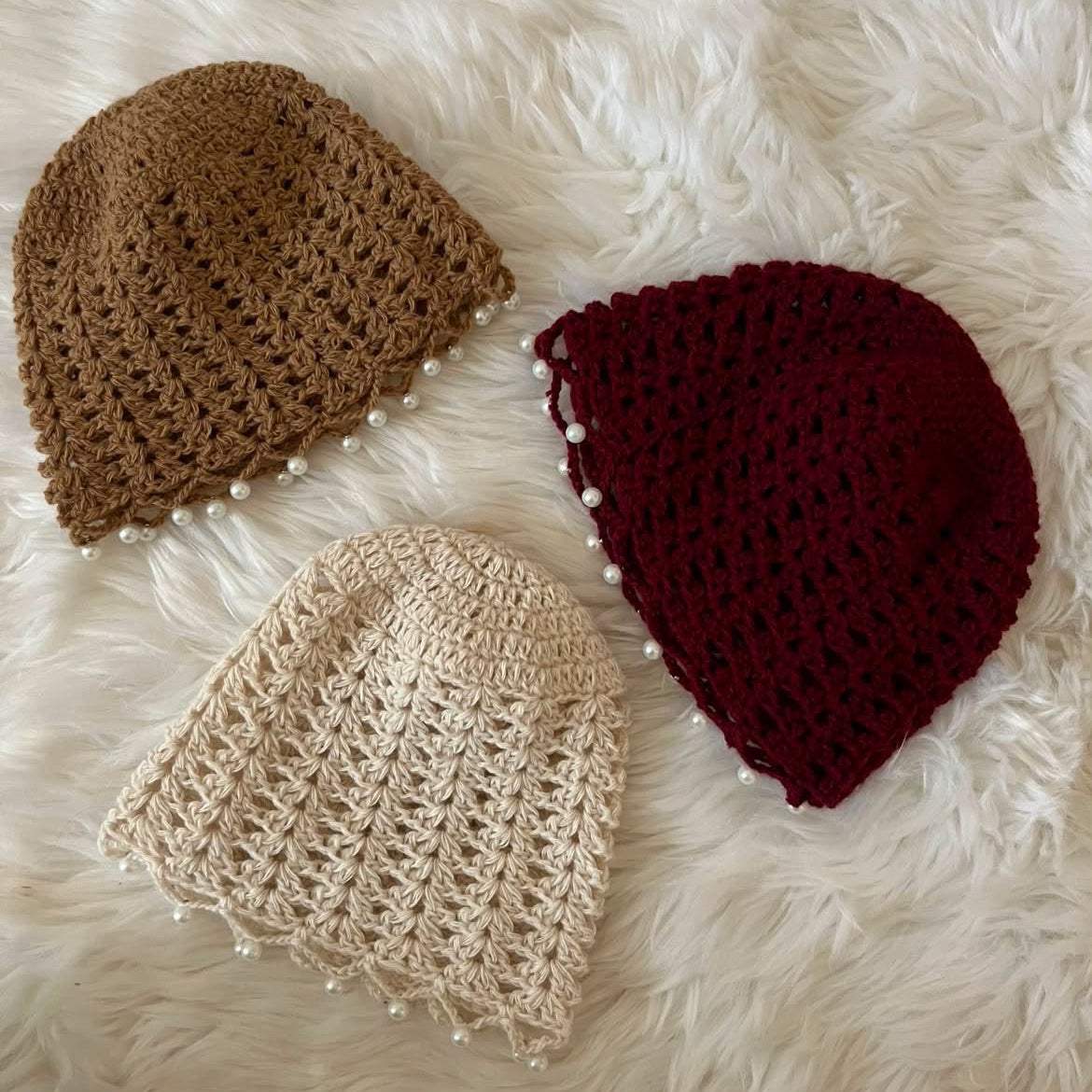 Three crocheted knit hats in brown, cream, and burgundy, each decorated with small white pearl beads along the edge, arranged on a white faux fur background.