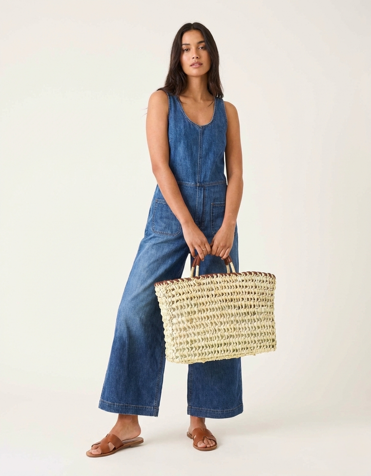 Woman wearing a blue denim jumpsuit holding a straw bag on a white background