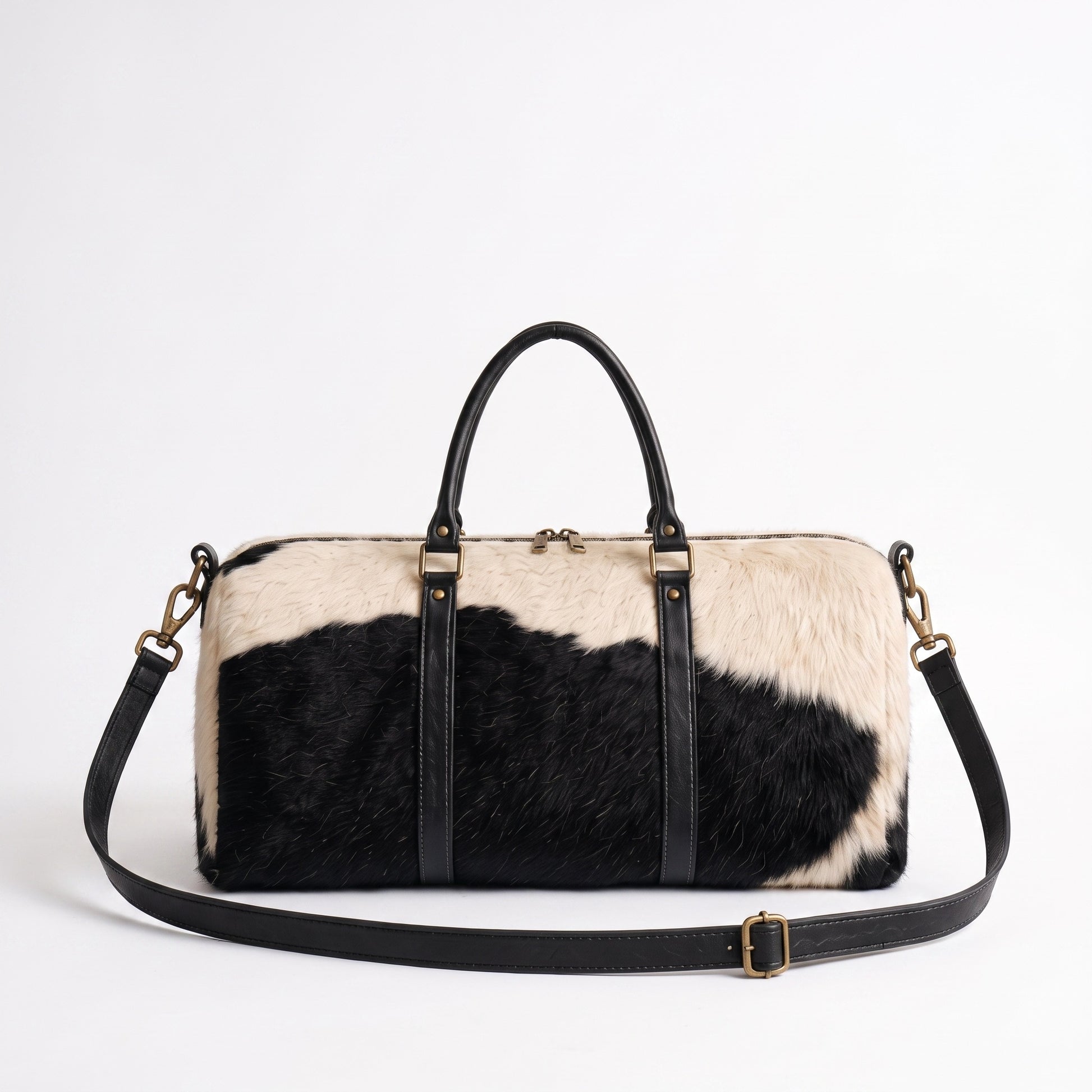 A duffle bag with a black and cream cowhide pattern, black leather handles and shoulder strap, and gold-tone hardware, photographed on a clean white background.