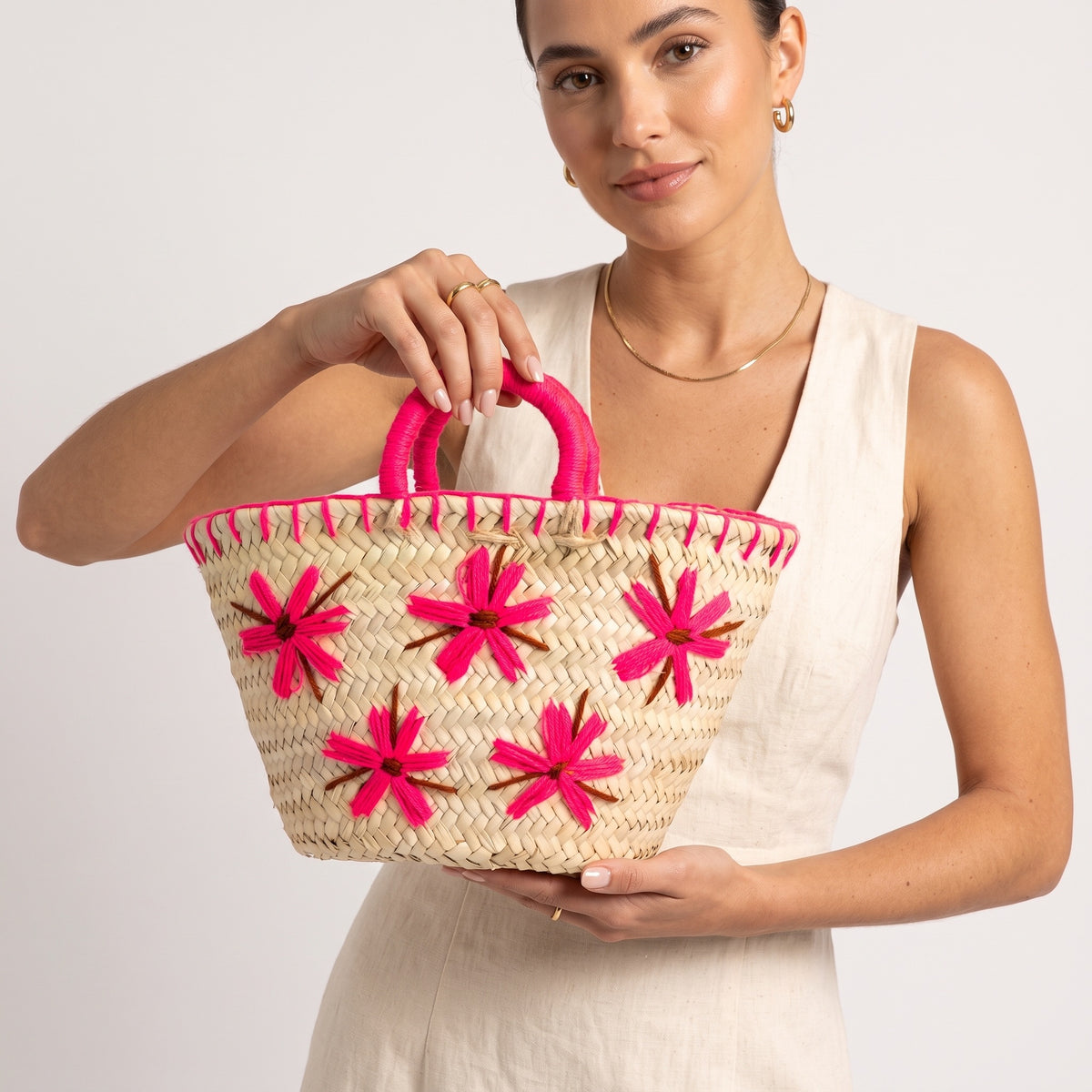 A woman in a white sleeveless dress holds a woven straw tote bag with bright pink handles and embroidered pink floral patterns on a light gray background.