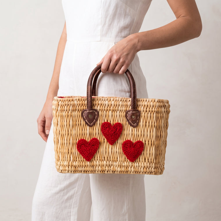A woman in a white dress holds a natural woven straw tote bag with dark brown handles and three red textured heart appliqués on the front, against a plain off-white background.