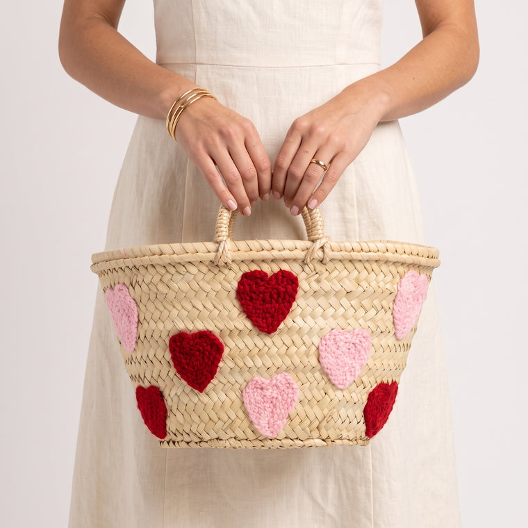 A woman in a cream linen dress holds a woven straw basket bag decorated with red and pink crocheted hearts, against a plain white background.