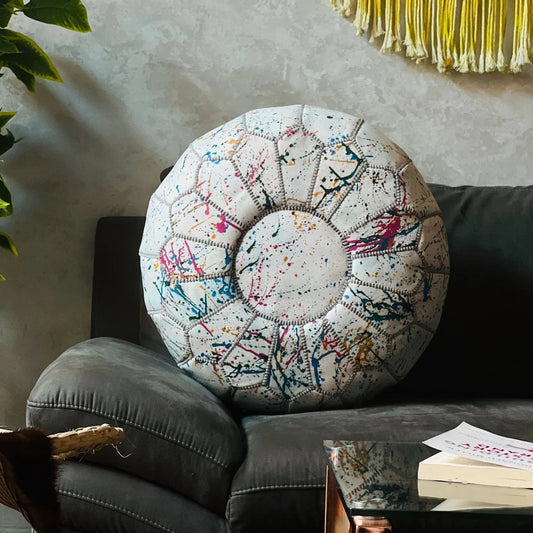 A round pouf ottoman with a colorful paint splatter pattern on a cream background, styled on a dark gray sofa in a living room setting.
