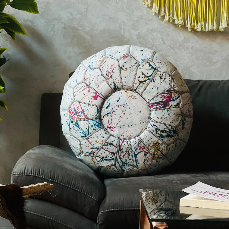 A round pouf ottoman with a colorful paint splatter pattern on a cream background, styled on a dark gray sofa in a living room setting.