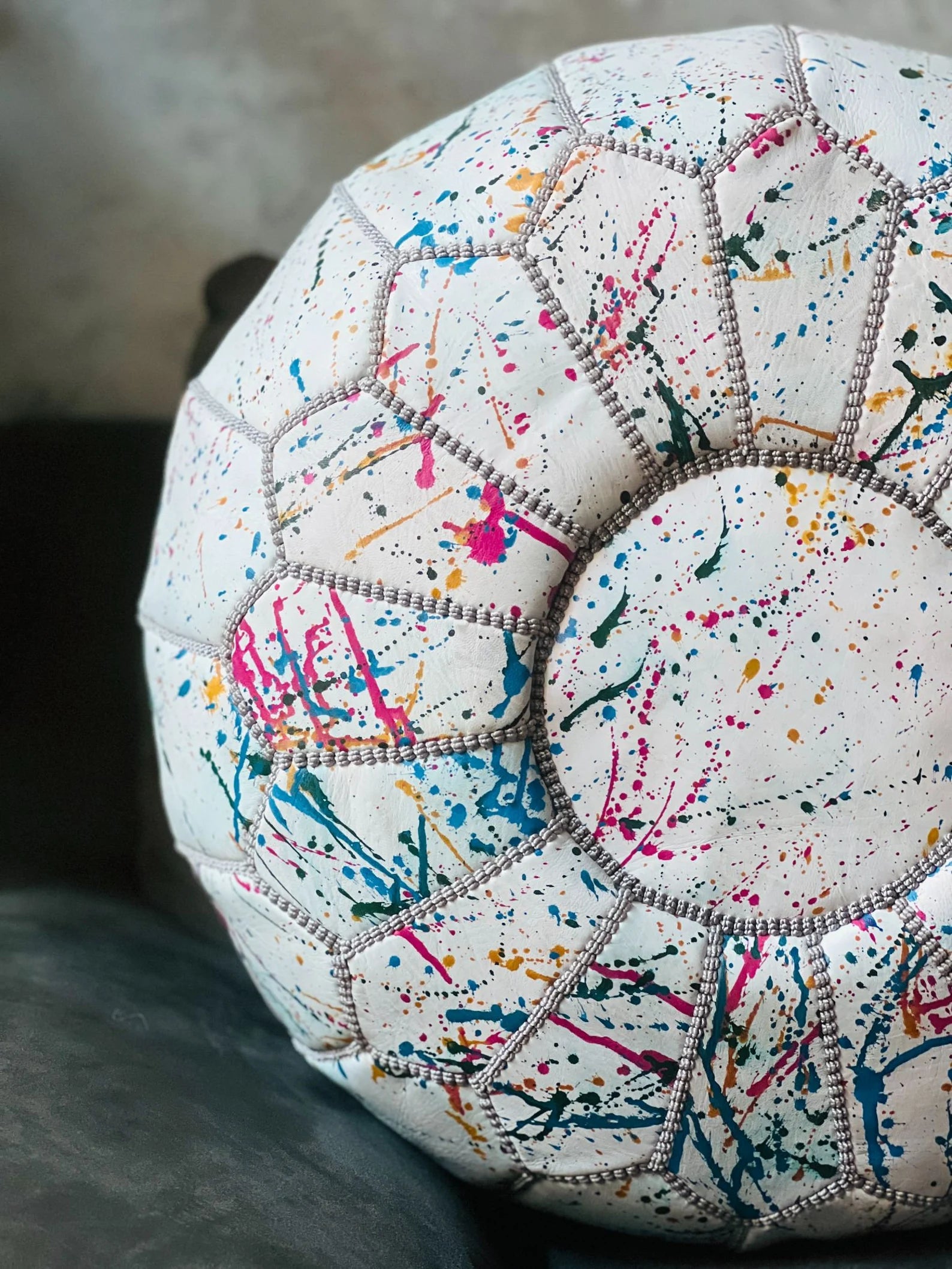 A close-up of a round pouf or cushion with a white base covered in colorful paint splatters and stitched geometric panels.